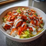 A close-up of a vibrant buffalo chicken bowl featuring shredded chicken, white rice, lettuce, carrots, and a drizzle of ranch dressing.