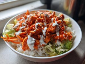 Close-up of a delicious buffalo chicken bowl featuring shredded orange chicken, white rice, lettuce, and a creamy ranch drizzle.