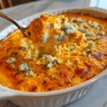 A spoonful of hot, cheesy Buffalo chicken dip topped with blue cheese crumbles being lifted from a white baking dish.