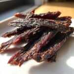 Close-up, sunlit photo of dark, seasoned venison jerky strips piled on a white plate.