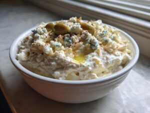A white bowl filled with creamy dirty martini dip, topped with blue cheese crumbles and green olives.