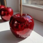 Close-up of a glistening, deep red candy apple with a wooden stick, sitting on a white surface near a window.