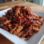 Close-up of tender, shredded slow cooker pulled pork heavily coated in rich, dark barbecue sauce on a white plate.