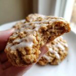 Close-up of a hand holding a soft iced oatmeal cookies broken in half, showing the moist interior and vanilla glaze.