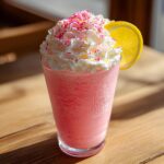 A tall glass of bright pink lemonade smoothie topped with whipped cream and colorful sprinkles.