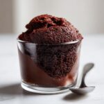 Close-up of rich, dark chocolate mousse with a fluffy top served in a small glass next to a spoon.
