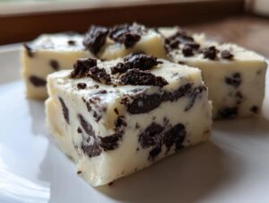 Three squares of creamy white Oreo fudge loaded with dark chocolate cookie pieces, sitting on a white plate.