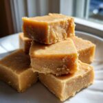 A stack of creamy, golden-brown peanut butter fudge squares sitting on a white plate near a window.