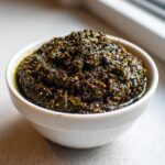 A close-up of a thick, dark green and brown olive dip, richly textured and oily, served in a small white bowl.