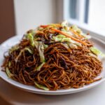 A large serving of dark, glossy fried noodles topped with fresh cabbage and carrot strips.