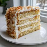 A tall slice of three-layer coconut cake with white frosting and toasted coconut flakes on the side.