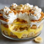 A scoop removed from a layered banana pudding trifle, showing vanilla custard, bananas, wafers, and whipped cream topping.