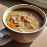 Close-up of a creamy white bean soup with chunks of ham and carrots, seasoned with pepper.