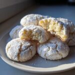 A stack of powdered sugar-dusted cool whip cookies on a light gray plate, one cookie broken open showing the soft interior.