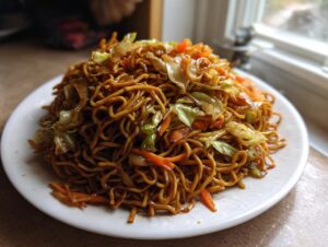 A generous serving of glossy, saucy fried noodles mixed with stir-fried cabbage and carrots on a white plate.
