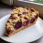 A close-up of a slice of plum cake featuring juicy purple plums and a golden crumble topping on a white plate.