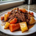 A plate of tender, shredded pot roast served with chunks of cooked carrots and potatoes in rich gravy.