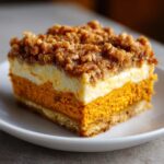 Close-up of a layered pumpkin crunch bar showing a crust, bright orange pumpkin layer, cream cheese layer, and crunchy topping.