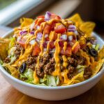A close-up of a hearty taco salad featuring seasoned ground beef, lettuce, diced tomatoes, red onions, black olives, and drizzled with bright orange cheese sauce, served with tortilla chips.
