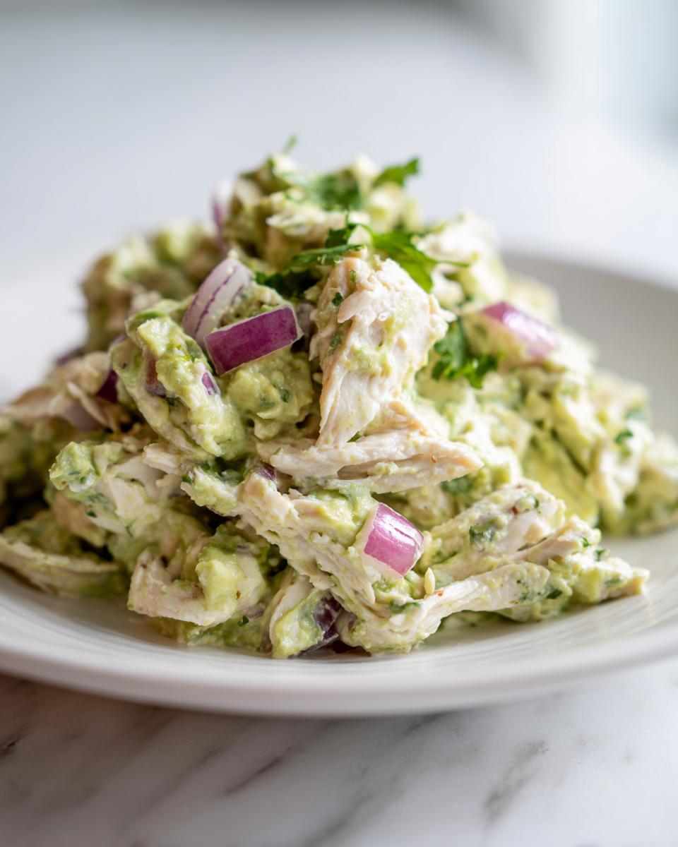 A close-up serving of creamy avocado chicken salad mixed with shredded chicken, chunks of red onion, and fresh cilantro.