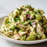 A close-up of creamy avocado chicken salad mixed with chunks of red onion and fresh cilantro on a white plate.