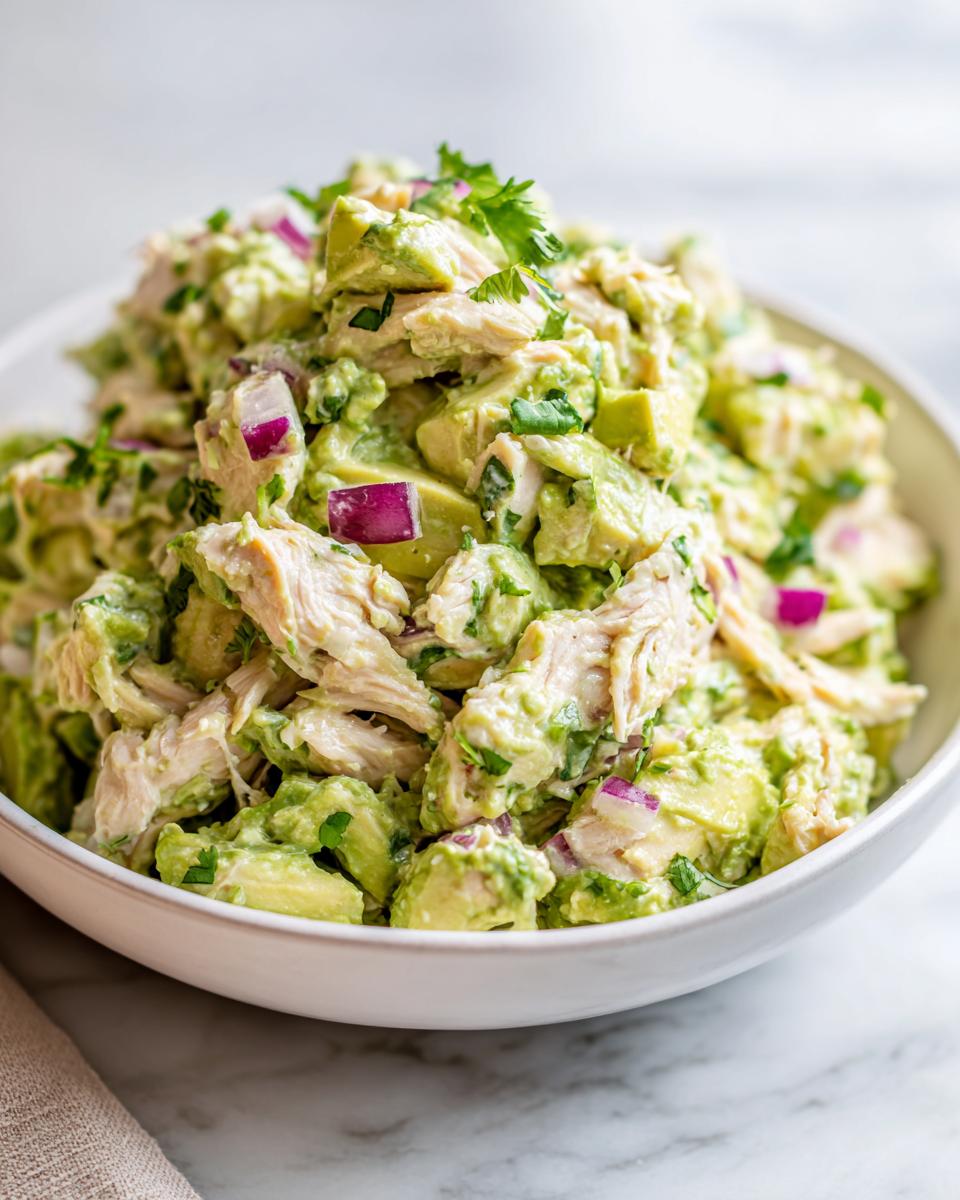 A close-up of creamy avocado chicken salad mixed with chunks of avocado, red onion, and cilantro, served in a white bowl.