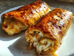 Two golden brown Baked Chicken Chimichangas on a white plate, one cut open showing stringy melted cheese and chicken filling.