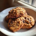A close-up stack of three golden brown banana oatmeal cookies with visible chocolate chips, served in a white bowl.