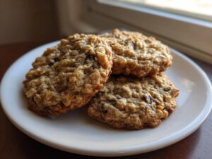 Three golden brown banana oatmeal cookies stacked slightly on a white plate near a window.