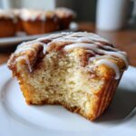 Close-up of a cinnamon roll muffin with a bite taken out, showing the soft interior and cinnamon swirl, topped with white icing.