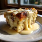 A close-up of a square slice of bread pudding recipe topped generously with rich vanilla sauce.
