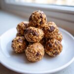 A stack of homemade chocolate chip protein balls made with oats, resting on a white plate near a window.
