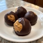 Four chocolate-dipped peanut butter balls on a white plate, one showing the creamy peanut butter filling.