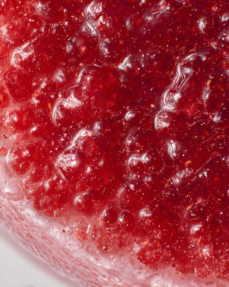 Extreme close-up texture shot of bright red, glistening strawberry freezer jam showing small fruit pieces and air bubbles.