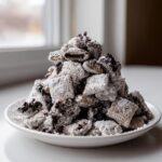 A tall pile of cookies and cream puppy chow coated in powdered sugar served on a white plate.