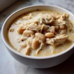 Close-up of a white bowl filled with creamy white bean and shredded chicken chili, topped with shredded cheese.