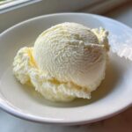A close-up shot of two scoops of creamy, pale yellow cottage cheese ice cream served in a white bowl.
