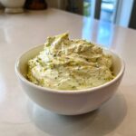 A close-up of creamy homemade pickle dip, flecked with dill and pickle pieces, served in a white bowl.