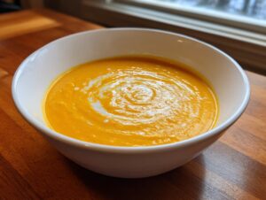 A close-up of a white bowl filled with vibrant orange, creamy pumpkin soup, swirled with cream.