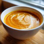 Close-up of a white bowl filled with creamy orange pumpkin soup, featuring a swirl of cream on top.