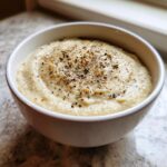 A close-up of creamy roasted cauliflower soup in a white bowl, seasoned heavily with black pepper.