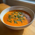 A close-up of a bowl of vibrant orange roasted tomato soup topped with fresh basil chiffonade and cracked black pepper.