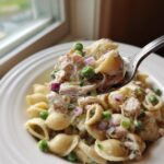 A spoonful of creamy tuna pasta salad featuring shell pasta, peas, and red onion being lifted from a white bowl.
