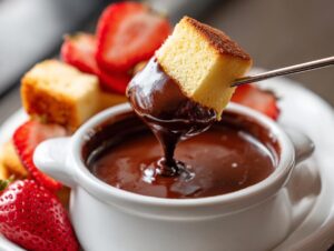 A piece of sponge cake is dipped into rich, melted chocolate fondue using a small fork, surrounded by fresh strawberries.