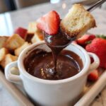 Close-up of cake and strawberry being dipped into rich chocolate fondue in a white ramekin.
