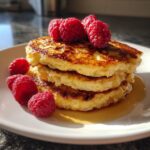 A stack of three golden brown almond flour pancakes topped with fresh raspberries and drizzled with syrup.