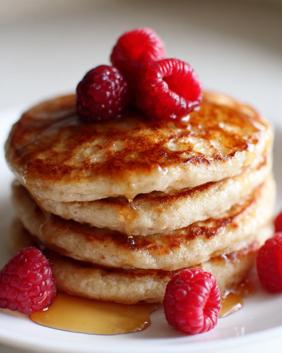 A tall stack of fluffy almond flour pancakes drizzled with syrup and topped with fresh raspberries.