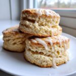 Close-up of three fluffy, layered scones stacked on a white plate, drizzled with white icing.