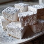 A stack of fluffy, white homemade marshmallows generously dusted with powdered sugar on a white plate.
