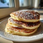 A stack of three fluffy oatmeal pancakes dusted generously with powdered sugar on a white plate.